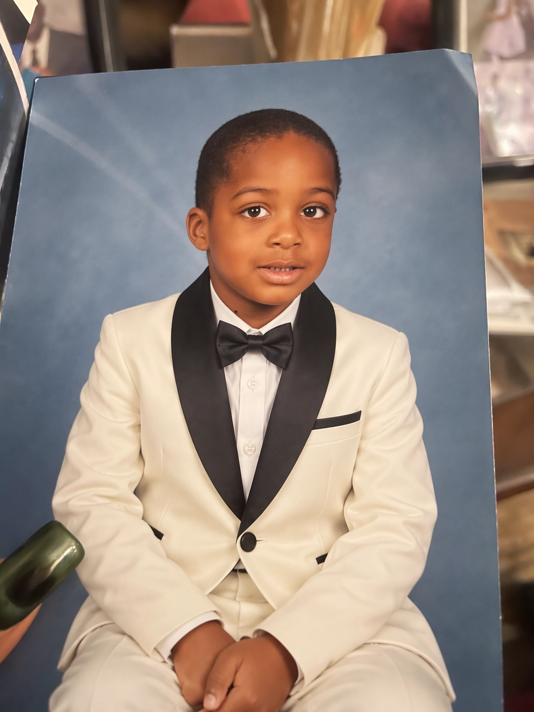 A printed studio portrait of a child, re-dressed by ChatGPT Images 2.5 into an ivory tuxedo with black lapels and a bow tie, keeping the original pose and blue background.